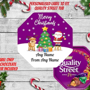 May include: A purple and white Christmas-themed label featuring a cartoon reindeer, Santa Claus, and a Christmas tree. The label reads "Merry Christmas" and "Any Name From Any Name". It is designed to fit a Quality Street tub.