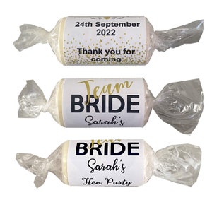 May include: Three white favor boxes wrapped in clear cellophane. The boxes are decorated with labels that say "Team Bride Sarah's" and "Thank you for coming 24th September 2022".
