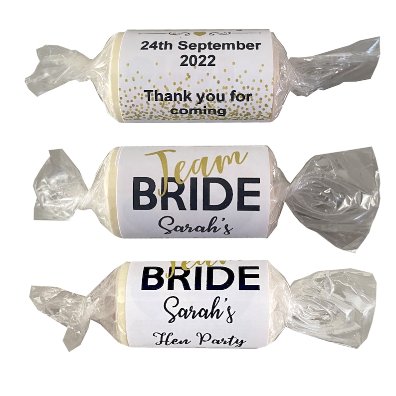 May include: Three white favor boxes wrapped in clear cellophane. The boxes are decorated with labels that say "Team Bride Sarah's" and "Thank you for coming 24th September 2022".