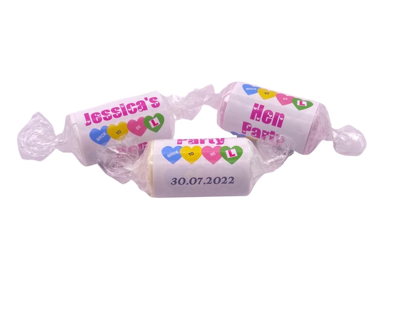 May include: Three pink and white wrapped candies with colorful hearts and the text "Jessica's Hen Party 30.07.2022" on one candy and "Hen Party" on another.