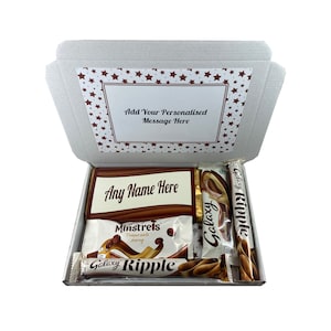 May include: A white cardboard gift box with a red star pattern on the lid. The box is open and contains a variety of chocolate bars, including Galaxy Ripple, Galaxy, and Minstrels. The box also includes a personalized message that says "Any Name Here".