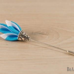 Puede incluir: Un prendedor de solapa decorativo con un diseño floral. Cuenta con pétalos de tela azul y blanca, una base de metal plateado y un alfiler largo y delgado. El prendedor se encuentra sobre un fondo claro de vetas de madera.