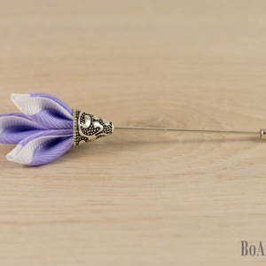 Op de afbeelding: Een revers speld in de vorm van een bloem met paarse en witte strepen en een sierlijke zilveren basis. De speld ligt op een lichtbruin houten oppervlak.