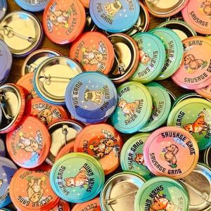 Pode incluir: Uma coleção de pinos redondos coloridos com várias raças de cães, incluindo Shiba Inus, Cocker Spaniels e Pugs. Os pinos apresentam ilustrações dos cães e texto que diz "Shiba Gang", "Cocker Gang" e "Pug Gang".