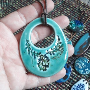 Könnte beinhalten: Ein blaugrüner Keramik-Anhänger mit floralem Design und einer braunen Lederschnur. Der Anhänger hat eine große ovale Form mit einem Kreis, der in der Mitte ausgeschnitten ist.