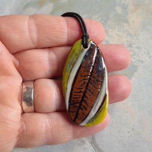 Puede incluir: Un colgante de cerámica con un diseño de hoja marrón, blanco y amarillo en un cordón negro.