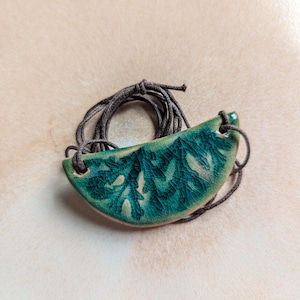 Puede incluir: Un colgante de cerámica verde y blanco con un diseño de hojas, colgado de un cordón marrón.