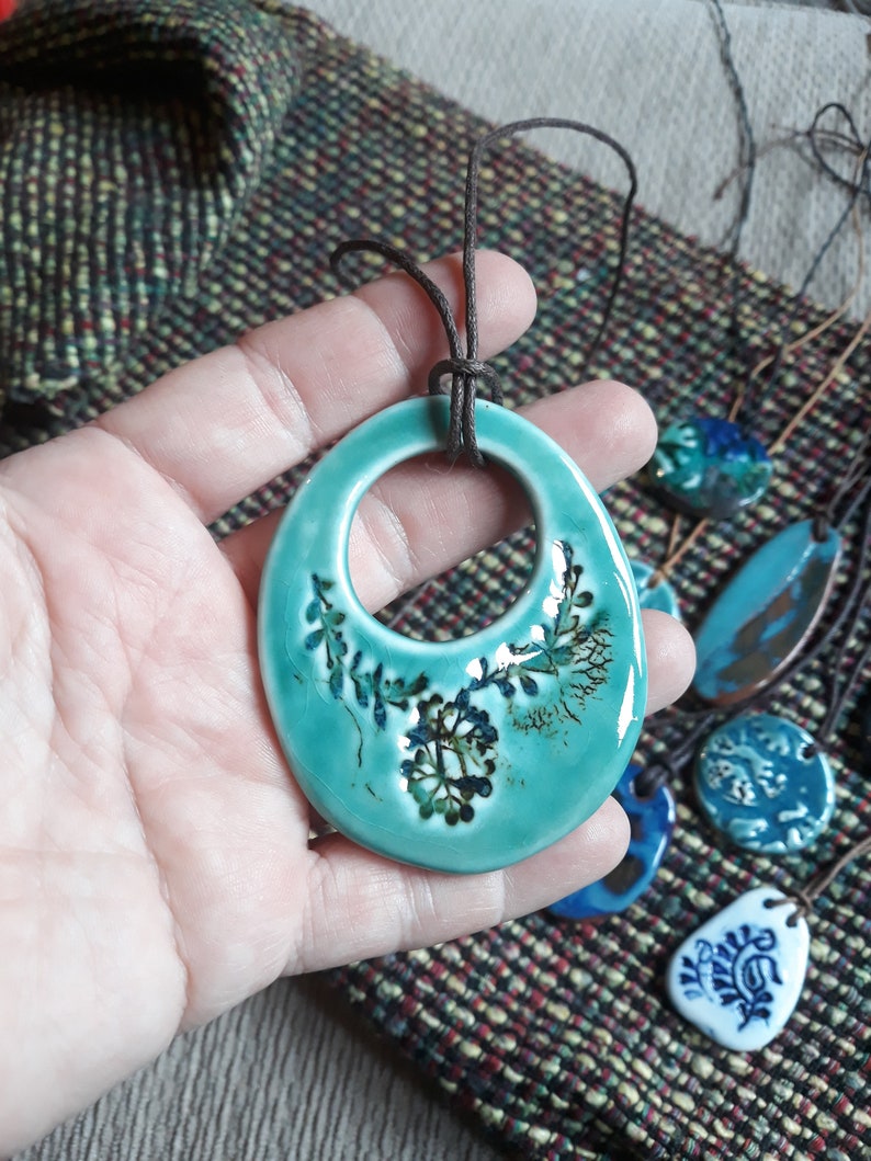May include: A teal blue ceramic pendant with a floral design and a brown leather cord. The pendant is a large oval shape with a circle cut out in the centre.