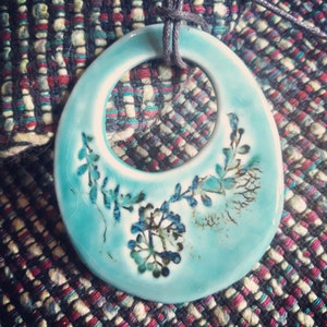 May include: A turquoise ceramic pendant with a floral design. The pendant has a ring in the centre and is attached to a black cord.