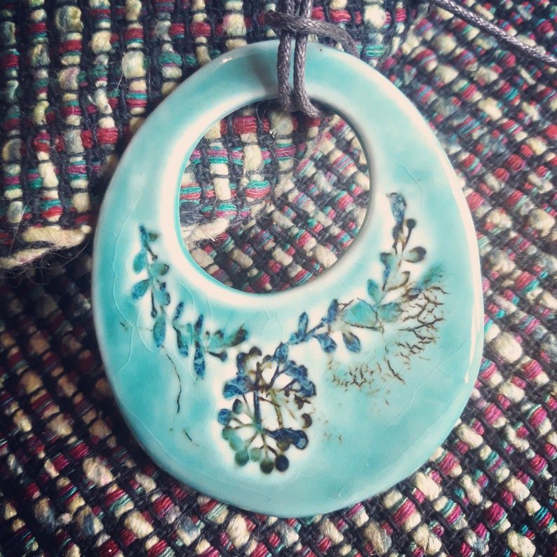 May include: A turquoise ceramic pendant with a floral design. The pendant has a ring in the centre and is attached to a black cord.