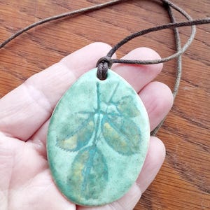 May include: A teardrop-shaped ceramic pendant featuring a glazed mint green surface with a dark green botanical leaf imprint. The pendant is suspended from a thin, dark brown cord that passes through a hole at the top of the ceramic piece.