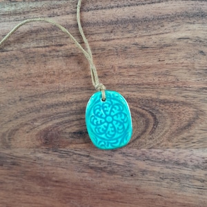May include: A turquoise ceramic pendant with a floral pattern, hanging from a beige cord. The pendant has an oval shape and a glossy finish. It rests on a wooden surface.
