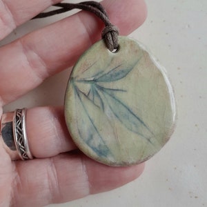 May include: A light green ceramic pendant with a delicate leaf design. The pendant is attached to a brown cord.