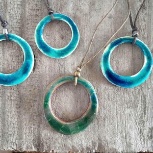 Puede incluir: Cuatro collares con colgantes de cerámica hechos a mano. Cada collar presenta un colgante circular con un esmalte brillante turquesa y verde. Los colgantes están ensartados en cordones de varios colores. El fondo es una superficie de madera envejecida.
