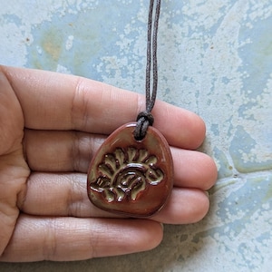 May include: A brown ceramic pendant with a green design of a stylised sun or flower. The pendant is on a brown cord necklace.