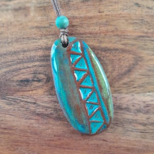 May include: A turquoise and brown ceramic pendant necklace. The pendant is an oval shape with a geometric design of triangles. The necklace has a brown cord and a turquoise bead.