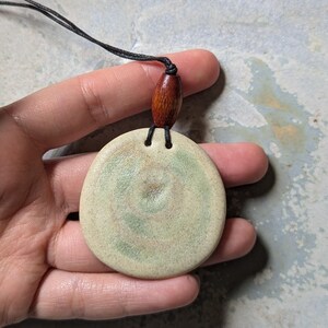 May include: A round, off-white ceramic pendant with a green and brown swirl design. The pendant is attached to a black cord with a brown wooden bead.