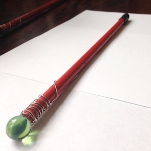 Pode incluir: Uma varinha vermelha artesanal com uma bolinha verde e amarela na base. A varinha é enrolada com arame prateado e tem uma ponta preta. A varinha tem aproximadamente 30 cm de comprimento.