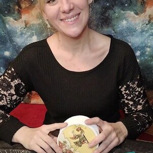 May include: A woman holds a tarot card with a yellow background and a depiction of a man holding a staff. The card is surrounded by a black surface with a crystal ball and a green box with a black and silver design.