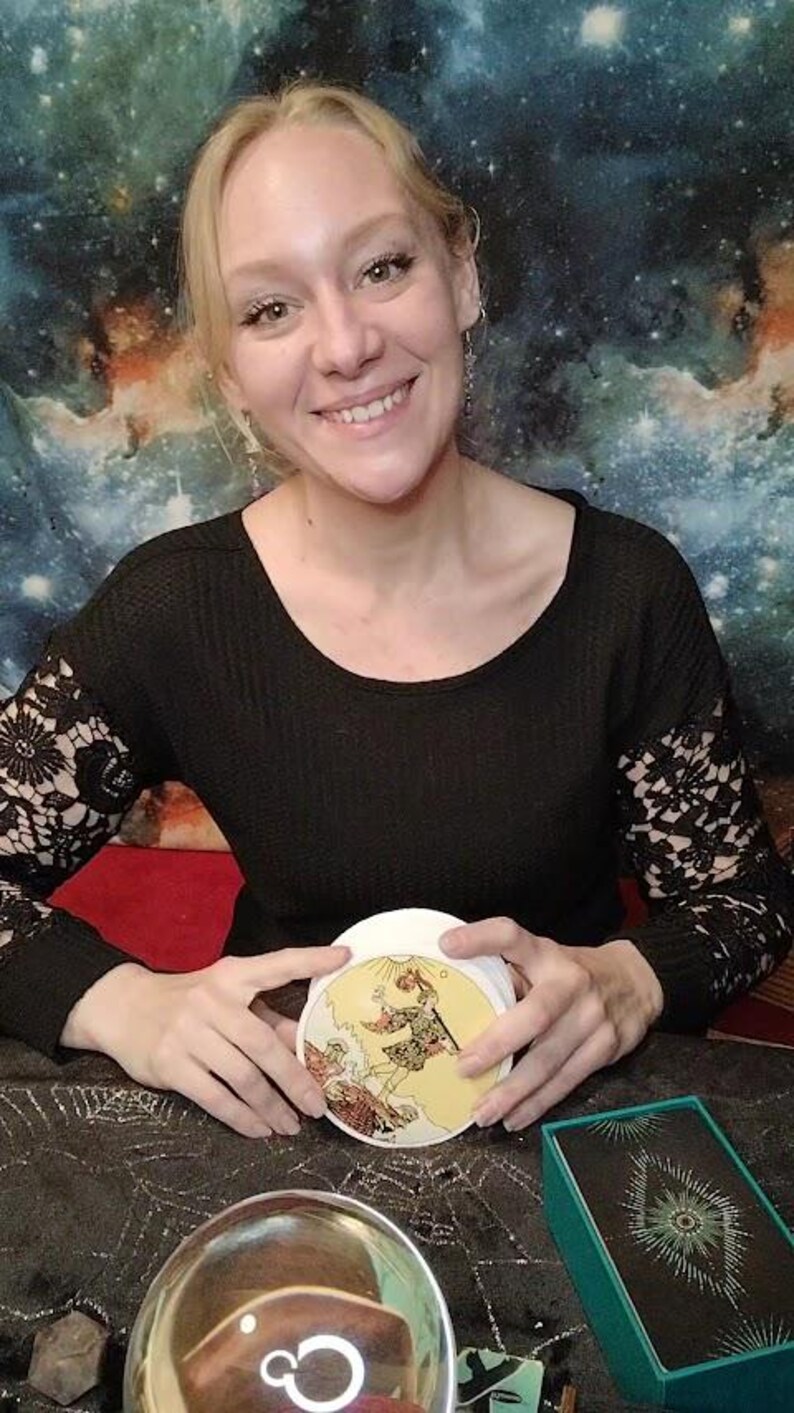 May include: A woman holds a tarot card with a yellow background and a depiction of a man holding a staff. The card is surrounded by a black surface with a crystal ball and a green box with a black and silver design.