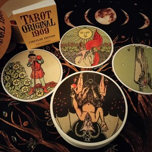 May include: Four tarot cards with circular white borders and colorful illustrations. The cards are laid out on a dark background with a pattern of branches and leaves. The cards depict the following: The Star, The Devil, The Hanged Man, and The Five of Pentacles. The cards are from the Tarot Original 1909 Circular Edition.