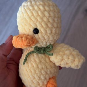 May include: A handmade, plush yellow duck toy with orange beak and feet, and a green bow. The duck has a black eye and is made of a soft, textured material. A cute, cuddly toy.
