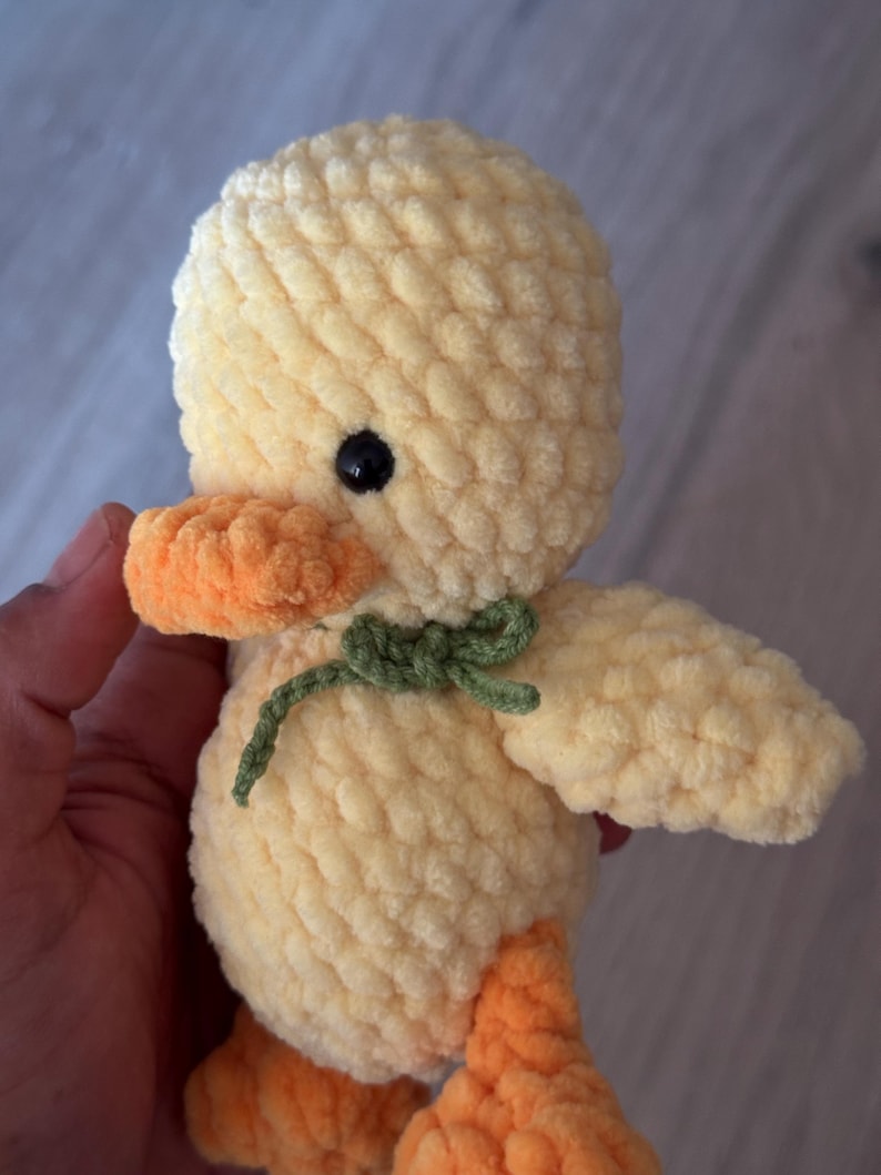 May include: A handmade, plush yellow duck toy with orange beak and feet, and a green bow. The duck has a black eye and is made of a soft, textured material. A cute, cuddly toy.