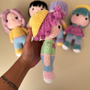 K&ouml;nnte beinhalten: Eine Sammlung handgefertigter H&auml;kelpuppen aus weichem, strukturiertem Garn. Die Puppen haben farbenfrohe Outfits, darunter gelbe H&uuml;te, rosa und lila Haare sowie pastellfarbene Kleidung. Jede Puppe hat ein rundes Gesicht mit schwarzen Augen und einem aufgestickten L&auml;cheln.