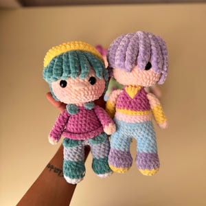 K&ouml;nnte beinhalten: Zwei handgefertigte H&auml;kelpuppen. Eine Puppe hat t&uuml;rkisfarbenes Haar und einen gelben Hut, tr&auml;gt ein rosa Hemd und eine gestreifte Hose. Die andere Puppe hat lila Haare, ein rosa-gelbes Oberteil und eine blaue Hose. Beide Puppen haben schwarze Augen.