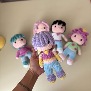 K&ouml;nnte beinhalten: Eine Sammlung handgefertigter H&auml;kelpuppen mit unterschiedlichen Haarfarben und Outfits. Die Puppen haben runde Gesichter, schwarze Augen und sind in farbenfrohe Kleidung gekleidet. Eine Puppe wird in einer Hand gehalten, um die weiche Textur und die Details zu zeigen.