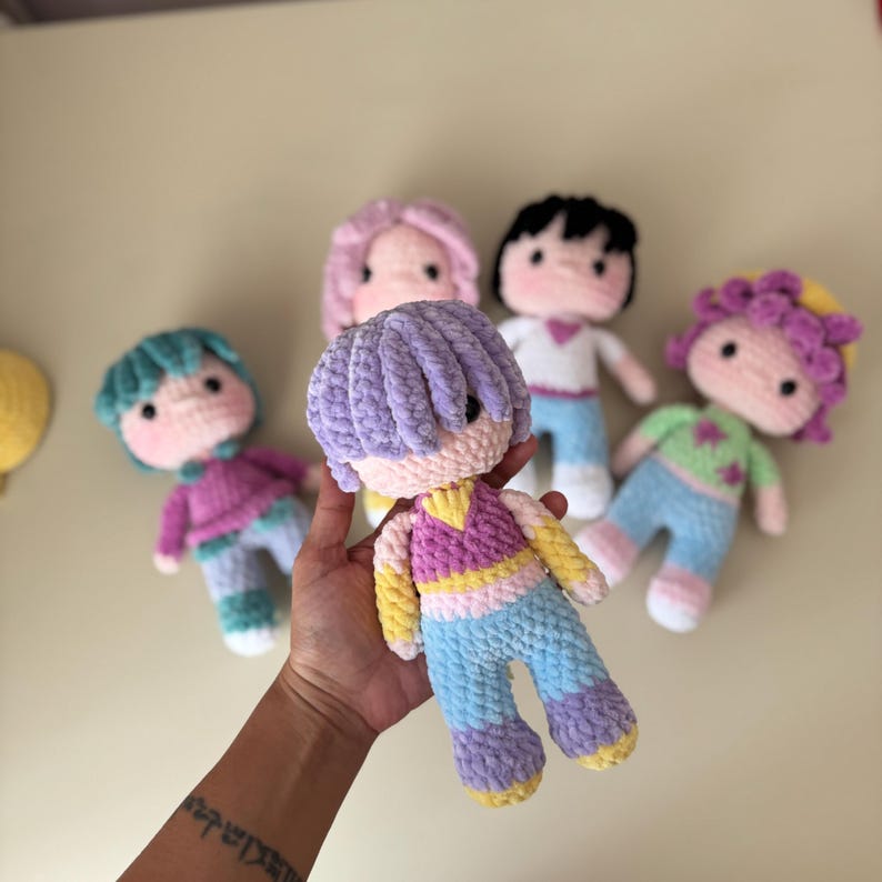 K&ouml;nnte beinhalten: Eine Sammlung handgefertigter H&auml;kelpuppen mit unterschiedlichen Haarfarben und Outfits. Die Puppen haben runde Gesichter, schwarze Augen und sind in farbenfrohe Kleidung gekleidet. Eine Puppe wird in einer Hand gehalten, um die weiche Textur und die Details zu zeigen.