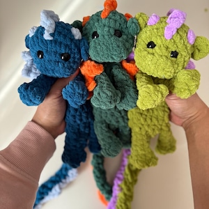 May include: Three handmade crochet dragon plushies in shades of blue, green, and lime green. Each dragon has a unique color scheme, with orange and purple accents. The dragons have black eyes and long, braided tails.
