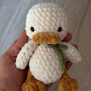 May include: A handmade, white crochet duck with a yellow-orange beak and feet. The duck has small black eyes and a green bow tied around its neck. The plush toy is soft and fluffy, perfect for a child's room or nursery.