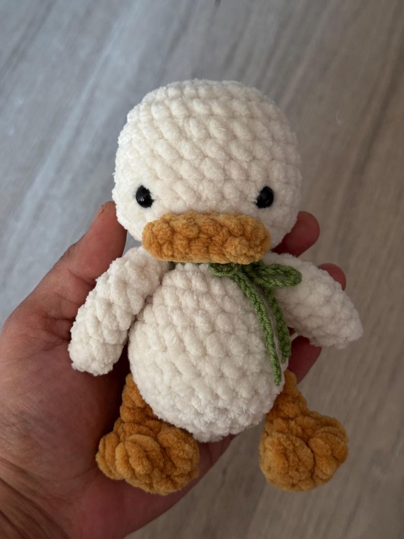 May include: A handmade, white crochet duck with a yellow-orange beak and feet. The duck has small black eyes and a green bow tied around its neck. The plush toy is soft and fluffy, perfect for a child's room or nursery.