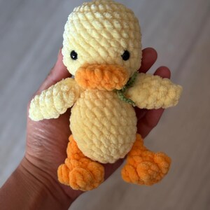 May include: A handmade, plush yellow duck with orange feet and beak, and black eyes. The duck has a green ribbon around its neck. The soft, textured toy is held in a person's hand.