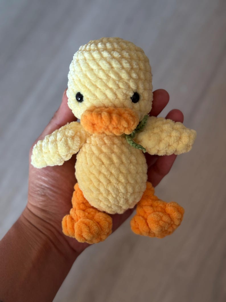 May include: A handmade, plush yellow duck with orange feet and beak, and black eyes. The duck has a green ribbon around its neck. The soft, textured toy is held in a person's hand.
