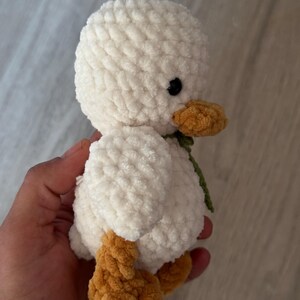 May include: A handmade, plush white duck with a textured, fluffy body. It features a yellow-orange beak and feet, and small black eyes. A green ribbon is tied around its neck. This soft toy is a charming, handcrafted item.