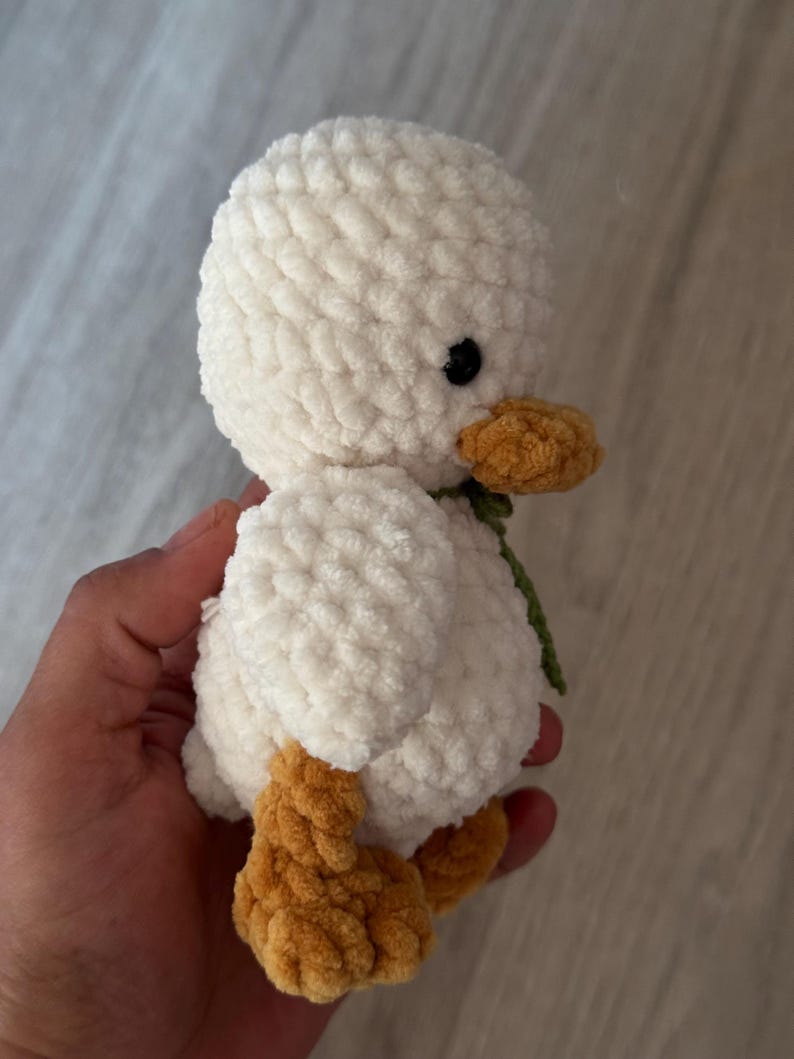 May include: A handmade, plush white duck with a textured, fluffy body. It features a yellow-orange beak and feet, and small black eyes. A green ribbon is tied around its neck. This soft toy is a charming, handcrafted item.