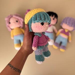 K&ouml;nnte beinhalten: Handgemachte H&auml;kelpuppe mit rosa Gesicht, blauen Haaren und gelbem Hut. Die Puppe tr&auml;gt eine rosa Jacke, blaue Hose und wei&szlig;e Schuhe. Im Hintergrund sind weitere Puppen mit unterschiedlich gef&auml;rbten Haaren und Outfits zu sehen.