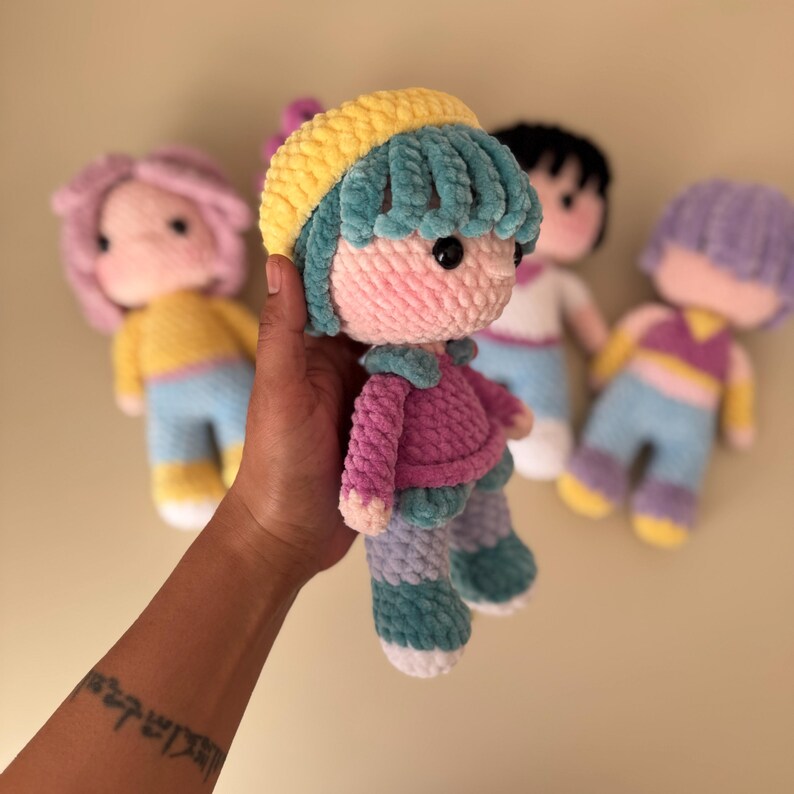 K&ouml;nnte beinhalten: Handgemachte H&auml;kelpuppe mit rosa Gesicht, blauen Haaren und gelbem Hut. Die Puppe tr&auml;gt eine rosa Jacke, blaue Hose und wei&szlig;e Schuhe. Im Hintergrund sind weitere Puppen mit unterschiedlich gef&auml;rbten Haaren und Outfits zu sehen.