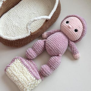 May include: A handmade crochet doll in shades of pink with a matching blanket and a brown and white crochet bassinet. The doll has a hooded head, black eyes, and is lying next to a small blanket. The bassinet is oval-shaped.