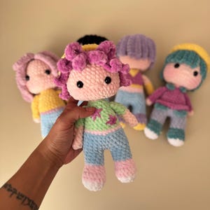 K&ouml;nnte beinhalten: Eine Sammlung handgefertigter H&auml;kelpuppen mit weichen, strukturierten K&ouml;rpern und farbenfrohen Outfits. Die Puppen haben rosa, lila und t&uuml;rkisfarbenes Haar mit passender Kleidung in Pastellfarben. Eine Puppe wird in der Hand gehalten.
