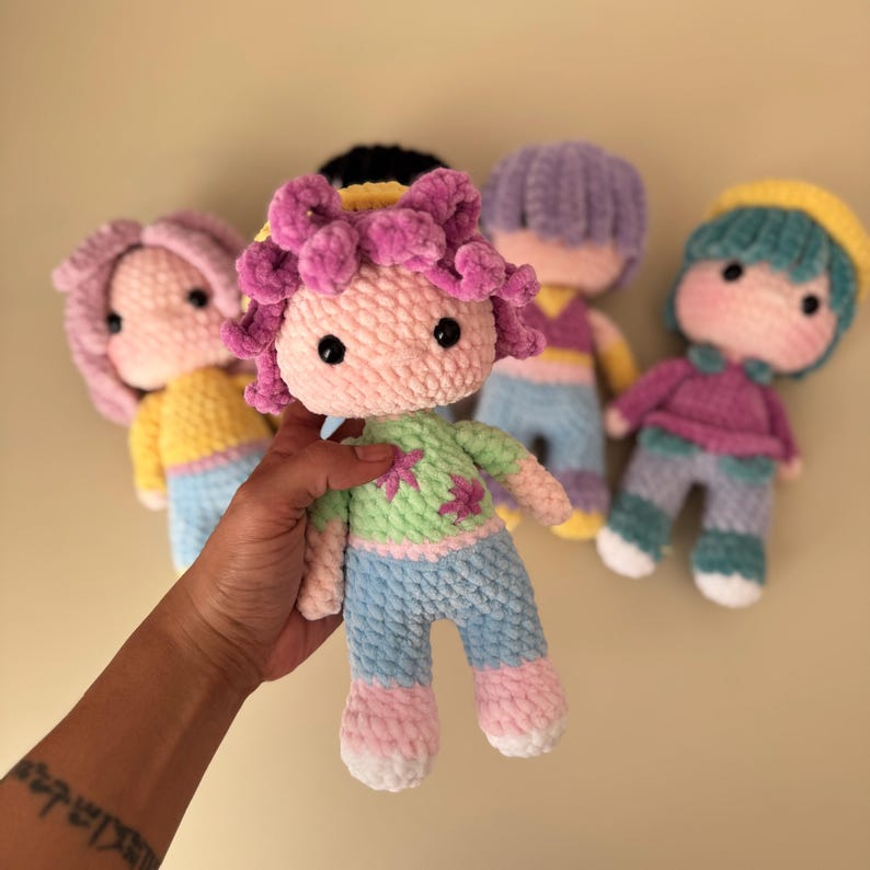 K&ouml;nnte beinhalten: Eine Sammlung handgefertigter H&auml;kelpuppen mit weichen, strukturierten K&ouml;rpern und farbenfrohen Outfits. Die Puppen haben rosa, lila und t&uuml;rkisfarbenes Haar mit passender Kleidung in Pastellfarben. Eine Puppe wird in der Hand gehalten.