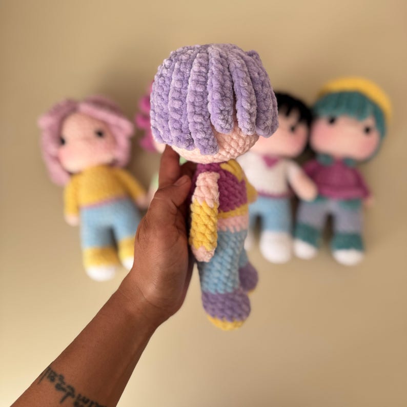 K&ouml;nnte beinhalten: Eine handgefertigte H&auml;kelpuppe mit lavendelfarbenem Haar, die ein farbenfrohes Outfit aus Gelb, Rosa, Lila und Blau tr&auml;gt. Im Hintergrund sind mehrere andere Puppen zu sehen, jede mit unterschiedlichen Haar- und Kleidungsfarben.