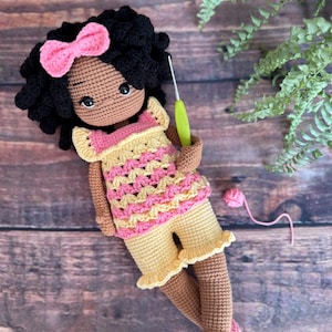 May include: Handmade crochet doll with black curly hair, a pink bow, and brown skin. The doll wears a yellow and pink top and shorts, and pink shoes. A crochet hook is held in its hand. The doll is on a wooden surface.