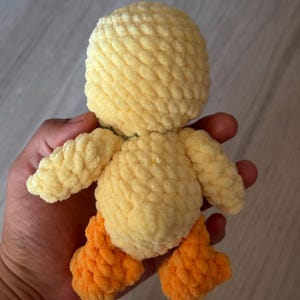 May include: A handmade, plush yellow duck with orange feet. The duck has a round head, small wings, and a soft, textured appearance. The toy is made of a fuzzy yarn material.