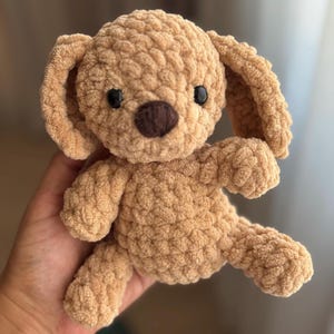 Può includere: Un cucciolo all'uncinetto fatto a mano, color marrone chiaro, con orecchie flosce, occhi neri e un naso marrone. Il peluche è morbido e strutturato, con cuciture visibili. Il cane è seduto, con le zampe e le gambe visibili.