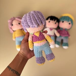 K&ouml;nnte beinhalten: Handgemachte H&auml;kelpuppe mit lila Frisur, tr&auml;gt ein rosa, gelbes und blaues Outfit. Andere Puppen mit unterschiedlichen Haar- und Kleiderfarben sind im Hintergrund. Die Puppe wird in einer Hand gehalten.