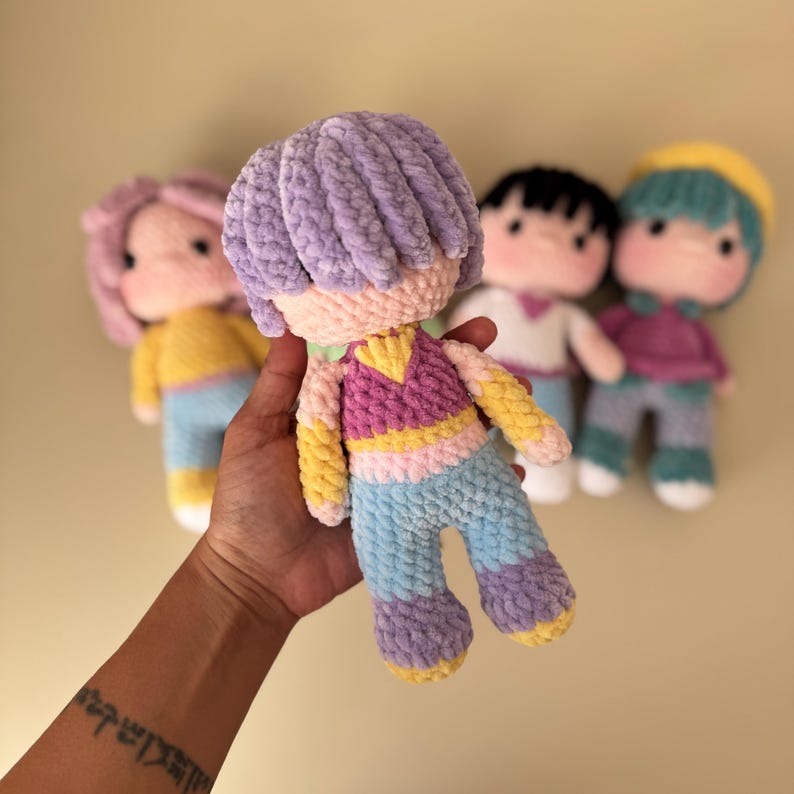 K&ouml;nnte beinhalten: Handgemachte H&auml;kelpuppe mit lila Frisur, tr&auml;gt ein rosa, gelbes und blaues Outfit. Andere Puppen mit unterschiedlichen Haar- und Kleiderfarben sind im Hintergrund. Die Puppe wird in einer Hand gehalten.