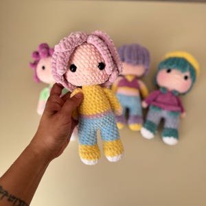 K&ouml;nnte beinhalten: Eine Sammlung handgefertigter H&auml;kelpuppen mit weichen, strukturierten K&ouml;rpern und Garnhaaren. Die Puppen haben eine Vielzahl von Farben, darunter Gelb, Blau, Rosa und Lila. Jede Puppe hat schwarze Knopfaugen und tr&auml;gt verschiedenfarbige Outfits.